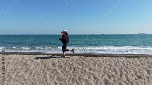 A person jogs on the sand by the ocean. Waves crash on the shore while boats are visible in the background. The sky is clear and blue, and it is daytime.