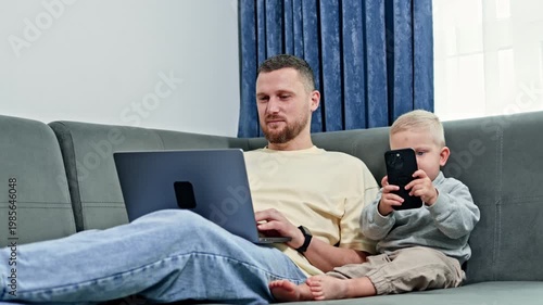 A man works on a laptop while a young child sits beside him holding a smartphone. They are relaxing together in a living room with curtains.