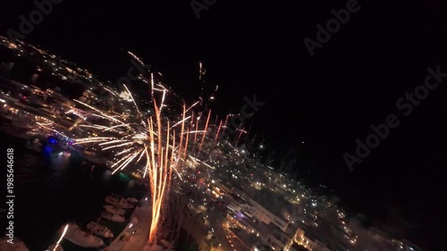 A cinematic FPV drone flight through colorful fireworks over a coastal town at night. Bright explosions illuminate the sky creating a festive and dynamic atmosphere