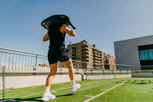 Young man performing functional training carrying a sandbag on shoulders in outdoor gym. Strength, endurance and fitness concept.