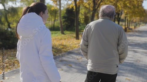 Elderly man and nurse walking in autumn park, back view of senior care