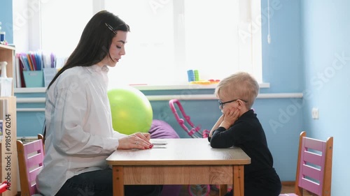 Preschool child interacts with teacher at a wooden table, focusing on educational materials in a vibrant classroom filled with toys and learning resources