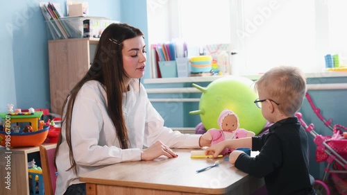 Preschool boy with glasses plays with a doll using a spoon and cup at a wooden table, with a teacher's hand visible in a colorful classroom
