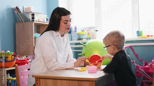 Preschool child interacts with teacher during playtime at a wooden table, surrounded by colorful toys and classroom elements in a bright indoor environment