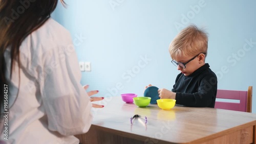 Preschool boy wearing glasses focuses on colorful bowls during a developmental activity at a wooden table, with teacher's hands in the foreground