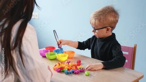 Young child wearing glasses engages in cutting colorful fruit pieces on a table, while a teacher is visible in the background assisting with the activity