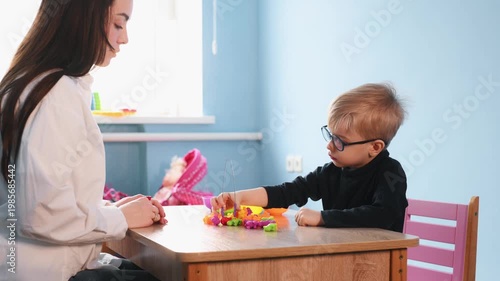 Preschool boy wearing glasses focuses on placing colorful shapes into a clear container on a wooden table, surrounded by bowls of various colors