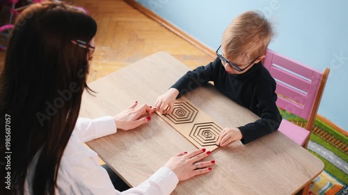 Young child wearing glasses focuses on a wooden maze activity at a table, while a teacher's hands assist in a bright classroom environment