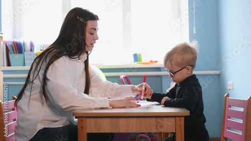 Young child wearing glasses uses markers to draw on paper with a teacher guiding at a wooden table in a vibrant classroom setting