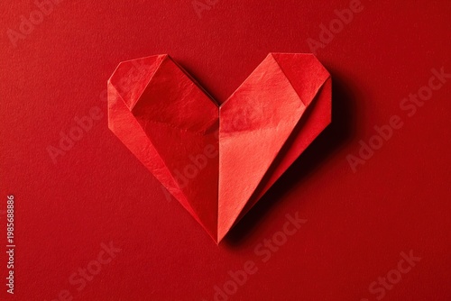 A red origami heart is situated on a bright red background. The focus is on the details of the folded paper. This setup shows a simple and clean arrangement for art