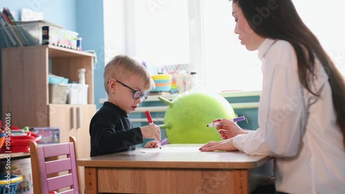 Preschool child works on a drawing with a teacher at a wooden table in a bright classroom, featuring educational toys and colorful decor