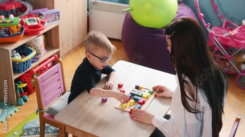 Preschool boy with glasses plays with colorful building blocks on a table while a teacher assists him in a vibrant classroom filled with educational toys