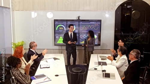 Diverse professionals in a boardroom applaud as two presenters share analytics on a large screen, showing dashboards and performance metrics during a corporate meeting