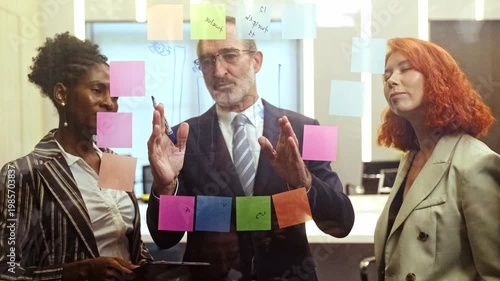 Team collaborating at an office glass wall, mapping ideas with colorful sticky notes as a leader explains strategy during a brainstorming meeting