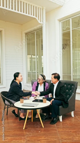 Three professionals meet in a bright lounge, shaking hands around a coffee table with documents and cups, signaling a successful agreement