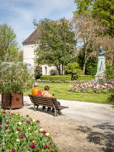 Elderly couple relax on bench in colourful French spring garden, enjoying peaceful sunny moment together.
