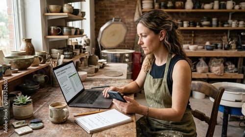 A female entrepreneur in a pottery studio working at a desk with a laptop and smartphone, reviewing small business funding options, warm earthy tones, clay tools and ceramic pieces visible in the