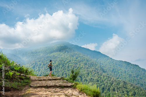 Bild auf Leinwand A person standing on a cliff overlooking a lush green mountain range with a blue