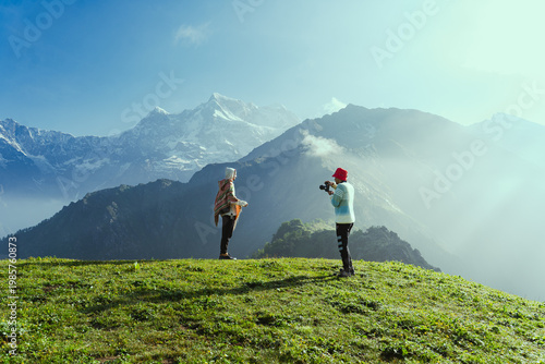 Photography Two hikers taking photos on a green hill with snow-capped mountains in the backg
