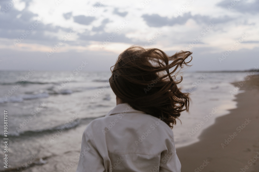 Obraz premium A cinematic rear view of a young woman standing on beach, her hair blowing wildly in the strong wind. The overcast sky and moody ocean waves create a feeling of peaceful solitude, raw nature, freedom