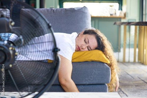 Woman sleeping on couch with fan during heatwave. Summer fatigue concept