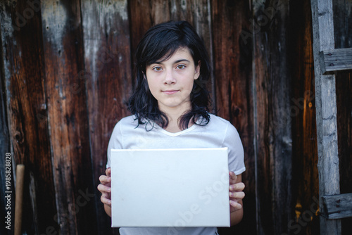 Young woman holding a blank white canvas in front of a wooden wall, minimal portrait with copy space.