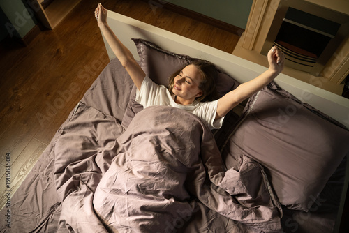 Happy young woman waking in bed, stretching arms overhead with eyes closed, smiling peacefully in a cozy bedroom, energized and enjoying a healthy morning routine