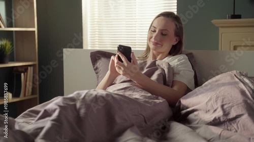 Happy young woman waking up in the morning and using her smartphone, scrolling through social media feeds, texting, and enjoying her leisure time before getting up from a comfortable bed
