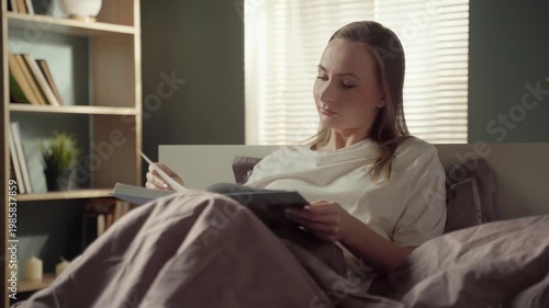 Peaceful young woman relaxing in her comfortable bed in the morning, enjoying a captivating book and turning pages in a cozy sunlit bedroom, representing a quiet lifestyle and hobby