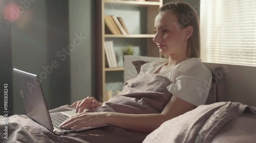 Smiling young woman comfortably sitting up in her bed and typing on a laptop, working remotely or chatting online in a cozy, sunlit bedroom during the morning or weekend