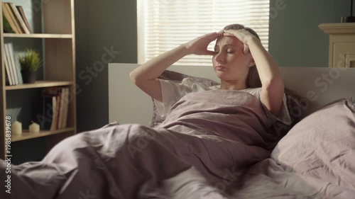 Young woman lying in bed suffering from a severe headache, massaging her temples with her fingers. Stressed and unwell person experiencing health problems and pain in the morning