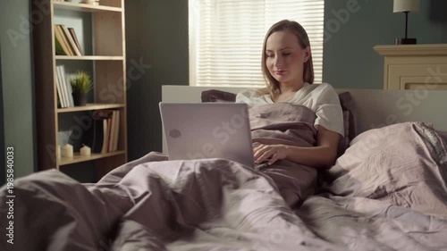 Relaxed young woman sitting comfortably in her bed under the duvet, smiling while working or browsing on her laptop computer early in the morning with soft sunlight coming through the window