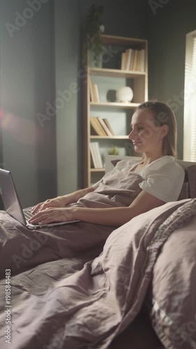Smiling young freelancer woman working remotely on her laptop while sitting comfortably in bed in the morning, enjoying a flexible work schedule and cozy home office environment