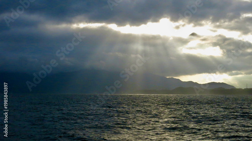 Sun rays breaking through storm clouds above dark tropical sea. Dramatic light beams illuminate distant mountains and coastline beneath moody overcast sky.