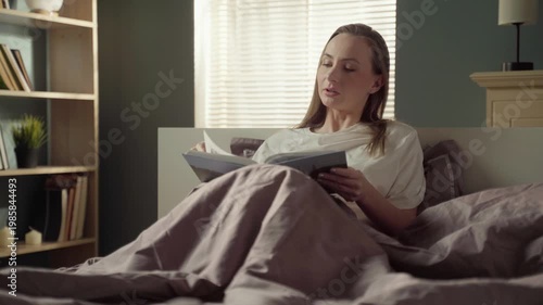 Relaxed young woman sitting in bed under the covers in the morning, smiling while looking through the pages of a family photo album and remembering happy moments of her life