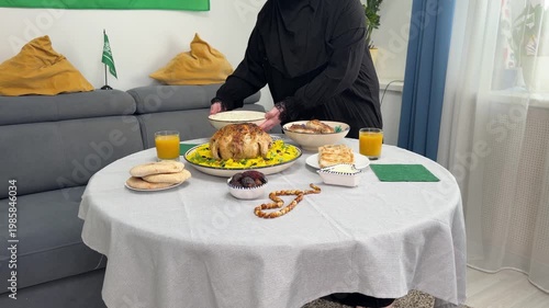 A woman in a black abaya sets table for a festive dinner. She serves a traditional Middle Eastern meal with roasted chicken, rice, dates, and bread. A Saudi Arabian flag is in background