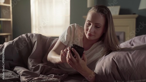 Cheerful young woman enjoying her morning routine, scrolling and texting on her smartphone while comfortably lying in bed, smiling and relaxing at home in her cozy sunlit bedroom