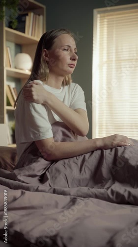 Young woman waking up in the morning and sitting on her bed, feeling intense shoulder pain and massaging the sore muscle after sleeping in an uncomfortable position