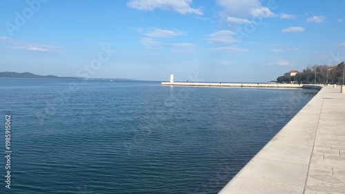 People walk along the path by the water while enjoying the view. The dock stands in the distance under a clear sky. Waves gently move along the shore.