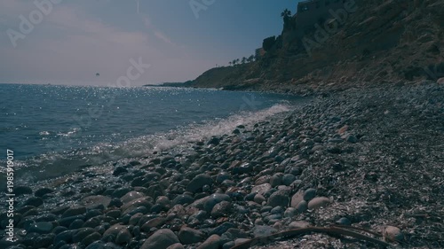 Waves hit the rocky shore in a coastal area during midday. Clear skies are visible above the water, showing the beauty of nature.