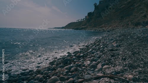 People explore a rocky beach. The waves crash against the stones. Sunlight glimmers on the water. The coastline stretches out with hills in the background.