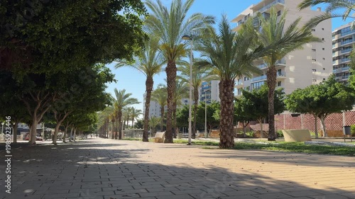 People walk along a path in a park that has palm trees and tall buildings. The weather is clear and sunny. The area looks lively and inviting for a stroll.