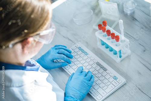 A laboratory professional wearing blue gloves types on a keyboard beside test tubes with red caps on a white table. The scene highlights modern scientific research and digital laboratory workflow