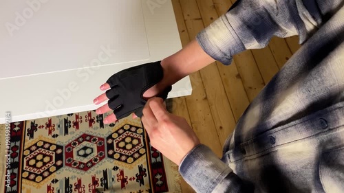 High-angle close-up video of a person putting on and fastening a black fingerless workout glove. Hands get ready for exercise, cycling, or weightlifting over a white table and wooden floor.