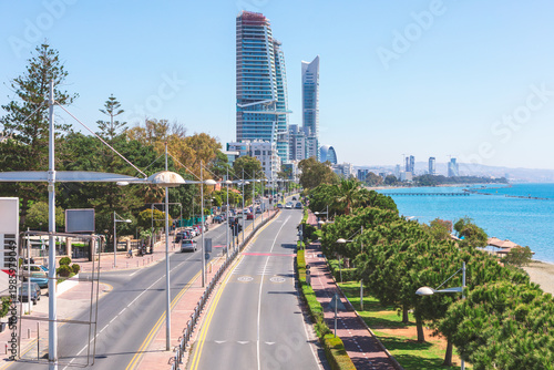 Coastal view captures the vibrant promenade of Limassol, Cyprus, lined with lush greenery and modern high-rises. Multi-lane road and dedicated cycling path run parallel to blue sea