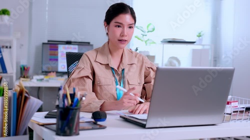 Focused Asian woman working intently on laptop at bright modern office desk during professional video communication reading corporate document for serious project data analysis