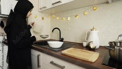 wide shot of a woman in hijab draining rice into a strainer over a white bowl in a bright home kitchen, documenting ingredient preparation for a festive family dish