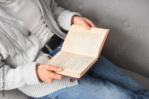 Young man reading book on grey sofa at home, closeup