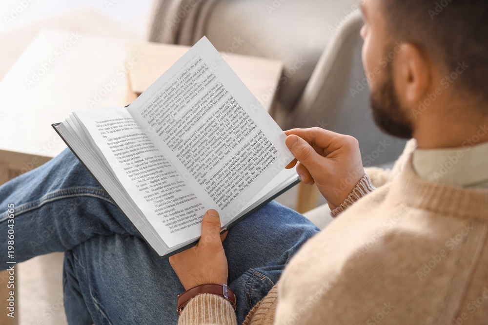 Naklejka premium Young man reading book on grey sofa at home, closeup
