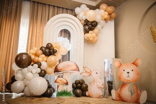 Beautifully decorated photo zone for a child's birthday party. Features a white arch, balloons in cream and brown tones, and large cutouts of a bear, bunny, and mushrooms in a forest theme.
